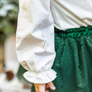 Détail d’une blouse blanche à manches longues avec petits nœuds argentés, assortie à une jupe verte scintillante, tenue de Noël pour fille de la collection Souris Mini.