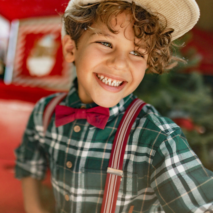 Petit garçon souriant portant une chemise verte à carreaux, des bretelles rouges et un nœud papillon, parfait pour un look chic des Fêtes.