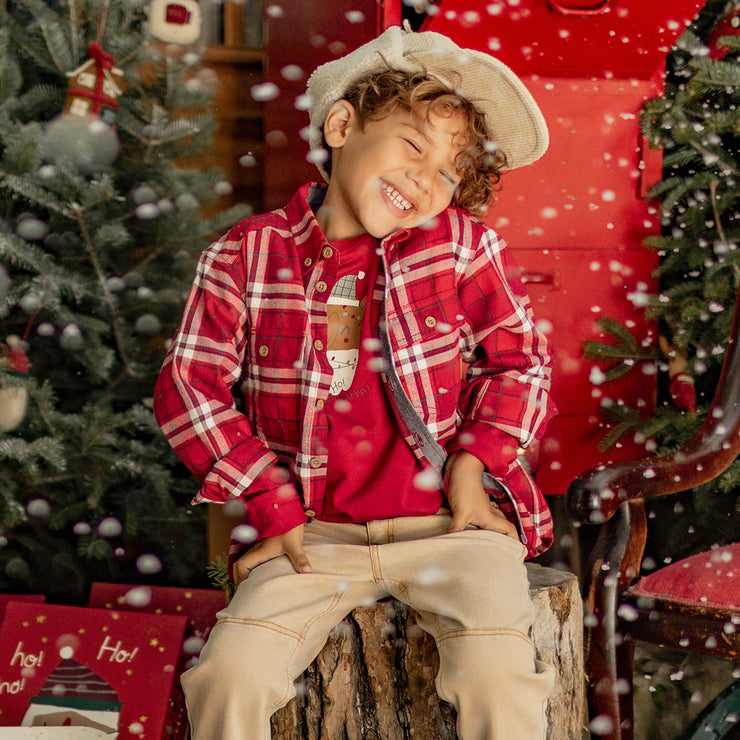 Petit garçon souriant portant une chemise à carreaux rouges et un t-shirt illustré d’un ours festif, assis dans un décor de Noël enneigé.
