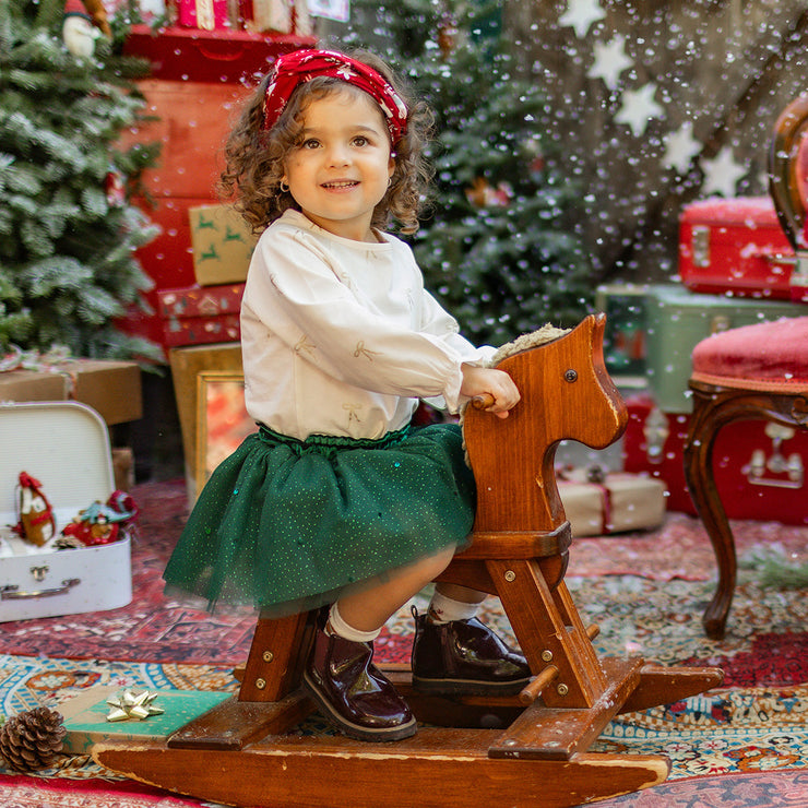Fillette souriante sur un cheval à bascule, portant une jupe verte à paillettes et un chandail crème, dans un décor festif de Noël, collection Souris Mini.