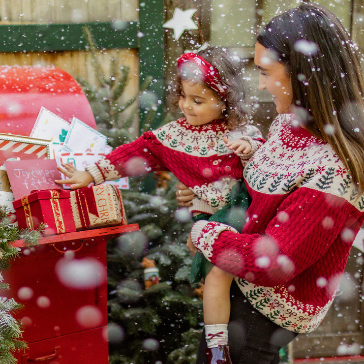 Maman et enfant portant des pulls rouges de Noël assortis en train de poster une carte de vœux, entourées de décorations festives, collection Souris Mini.