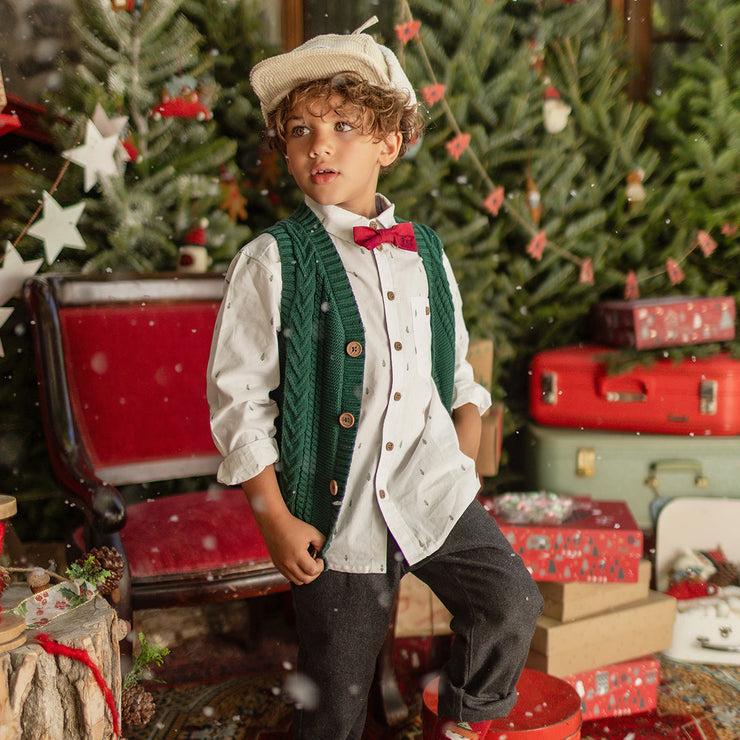 Petit garçon habillé pour Noël avec une chemise blanche à motifs, un nœud papillon rouge et un gilet vert en tricot, posant dans un décor festif avec sapins et cadeaux.